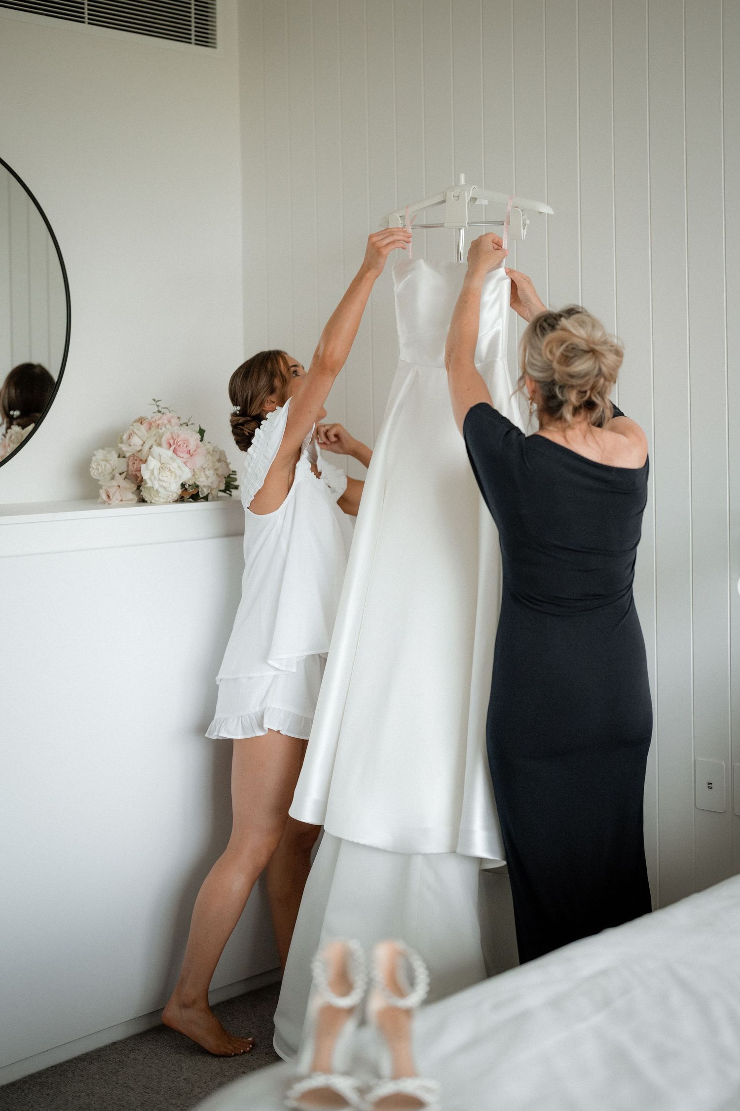 Two people carefully hang an elegant white wedding dress on a hanger in a bright room with white walls and bridal accessories.
