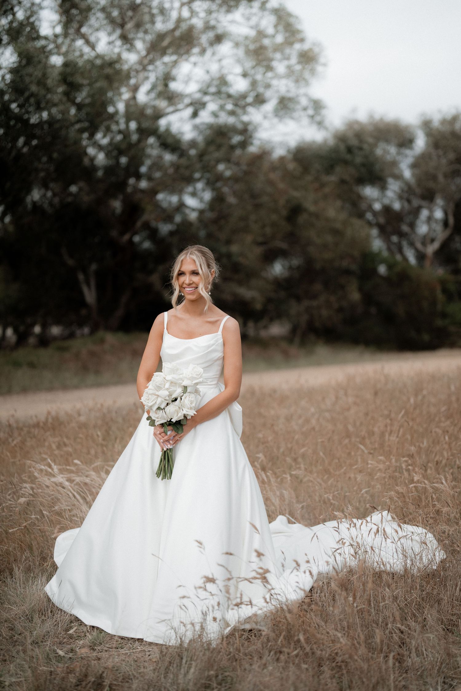 Elegant bride in flowing white gown holds bouquet while standing in rustic field at sunset.