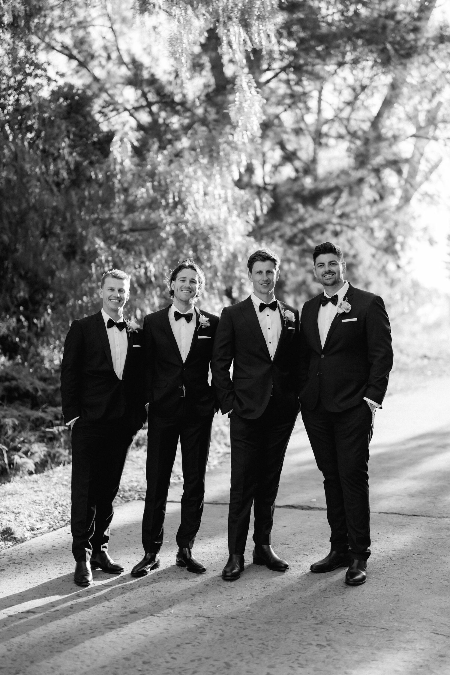 Groomsmen in black tuxedos and bow ties pose casually together on a tree-lined path in black and white photography.