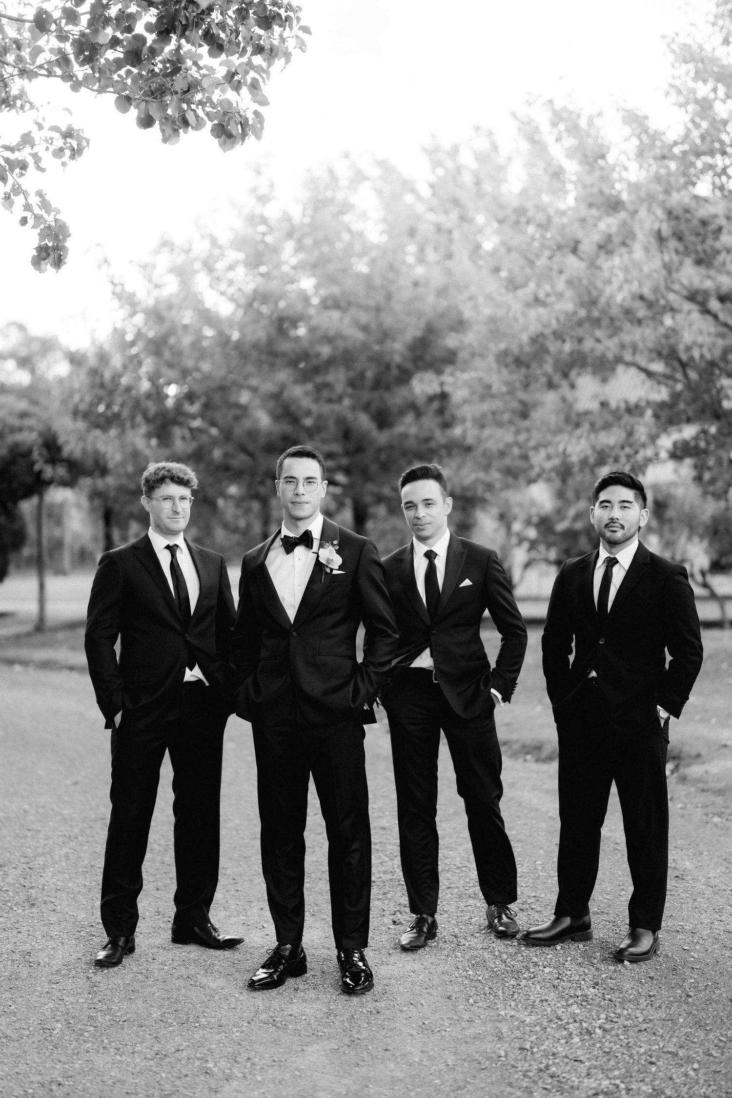 Groom and three groomsmen in black suits posing outdoors in black and white photo.