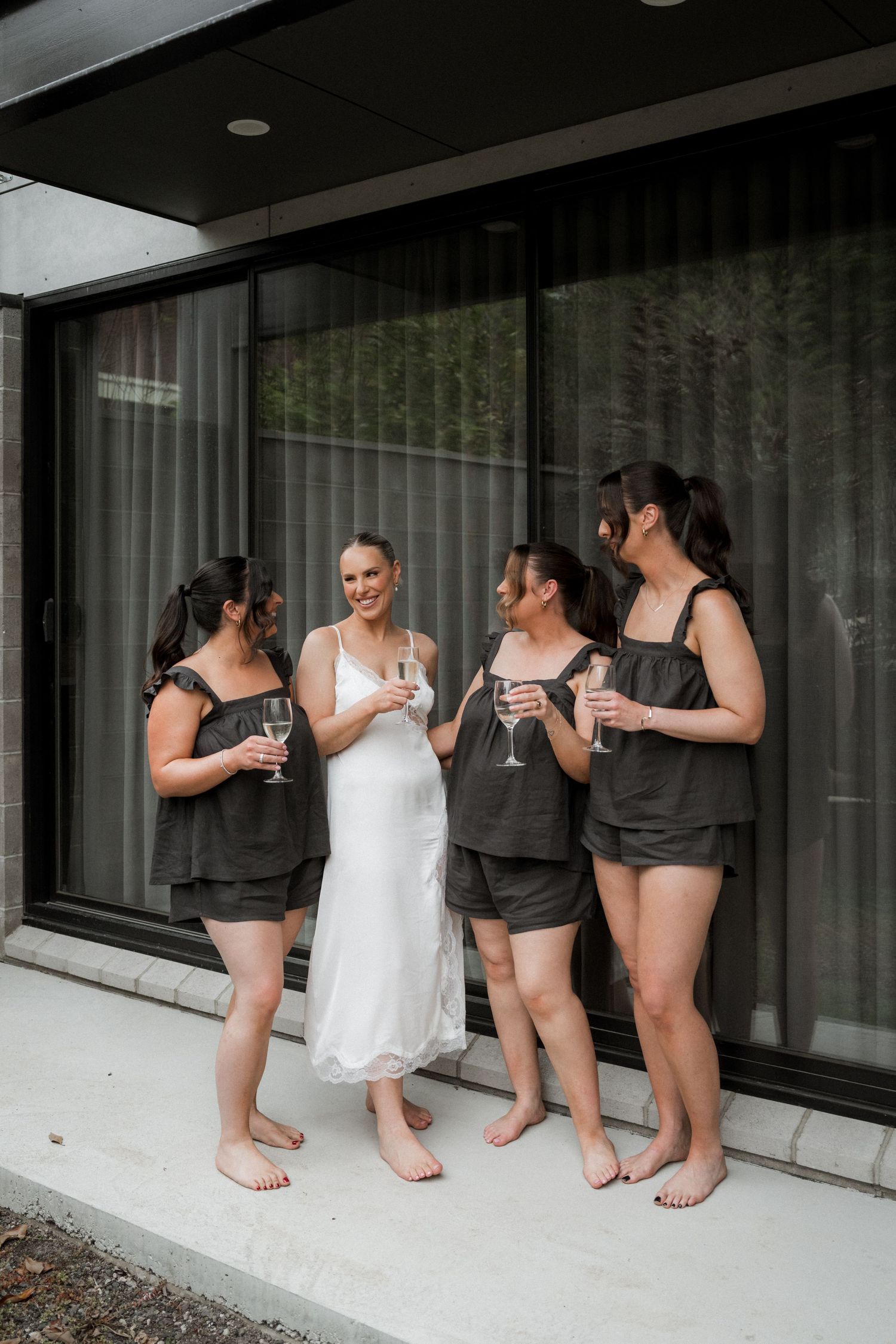 Bride in white slip dress with bridesmaids in dark pajamas toasting champagne on a modern patio.