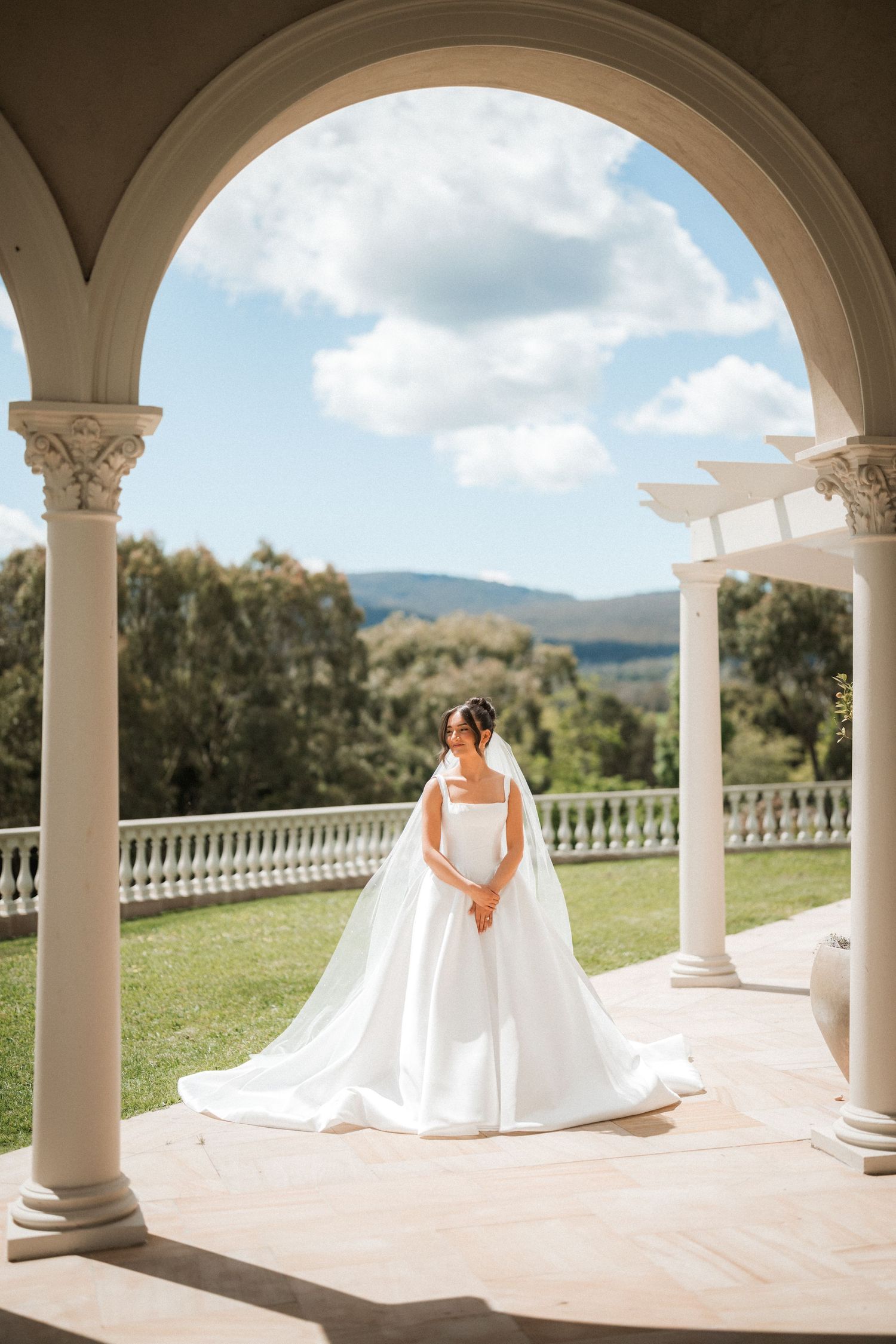 Bride in a white satin gown and long veil stands beneath a stone archway overlooking a green lawn and distant hills.