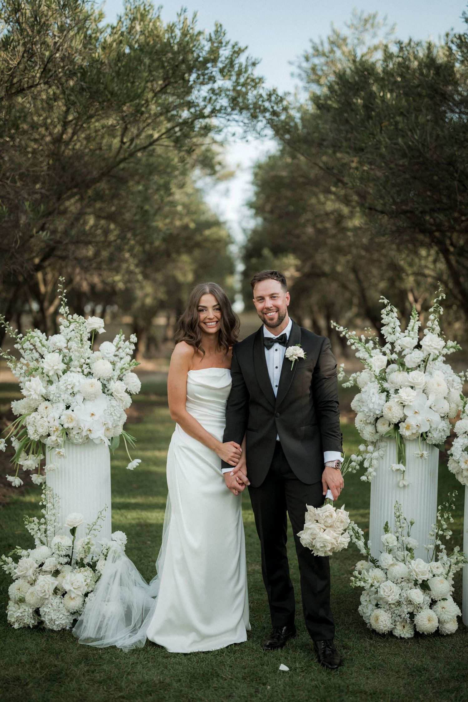 Elegant white floral arrangements frame a wedding couple in an outdoor garden setting.
