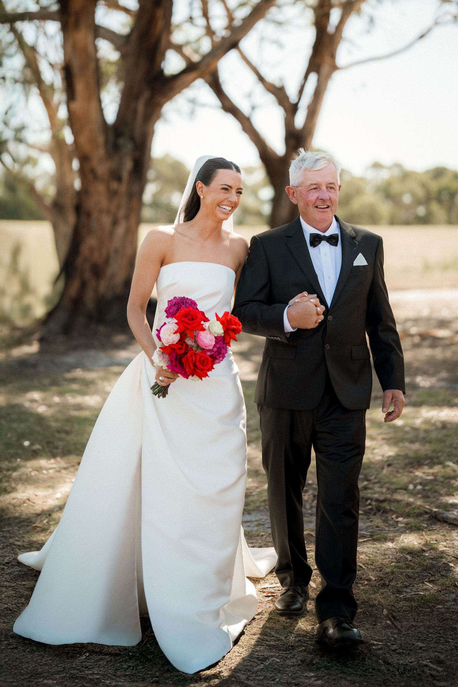 A bride in elegant white dress walks arm in arm with an escort under large trees.