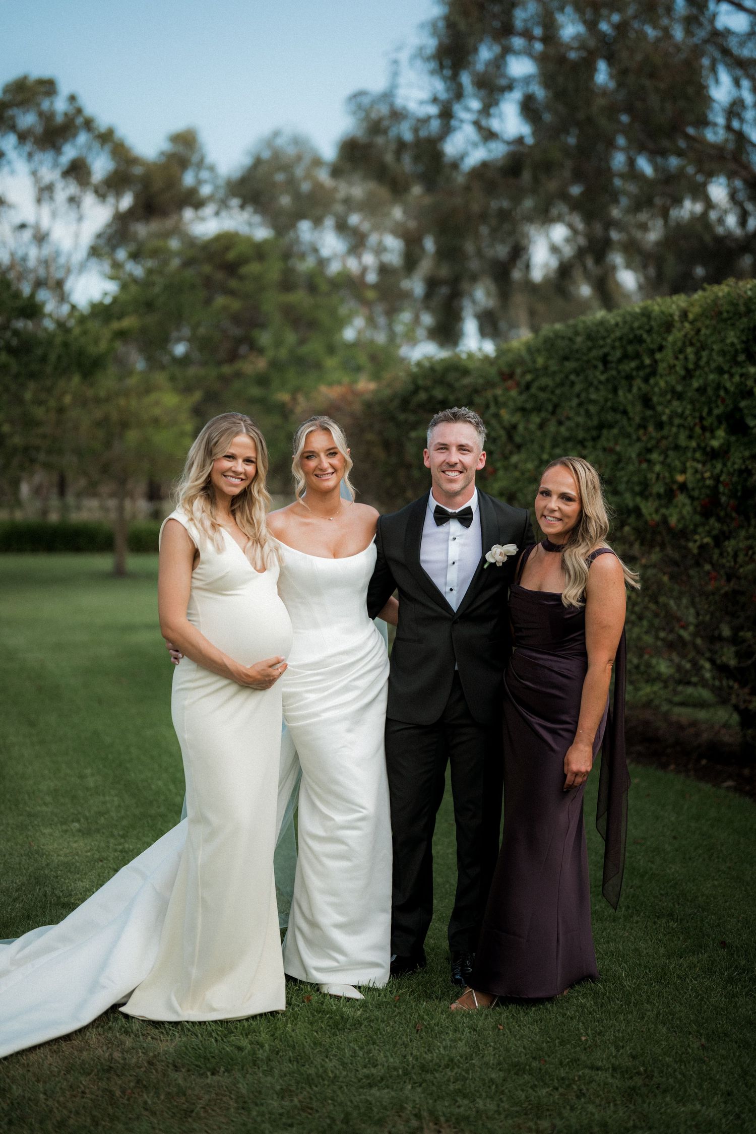 Wedding party poses together on grass with hedges in background.