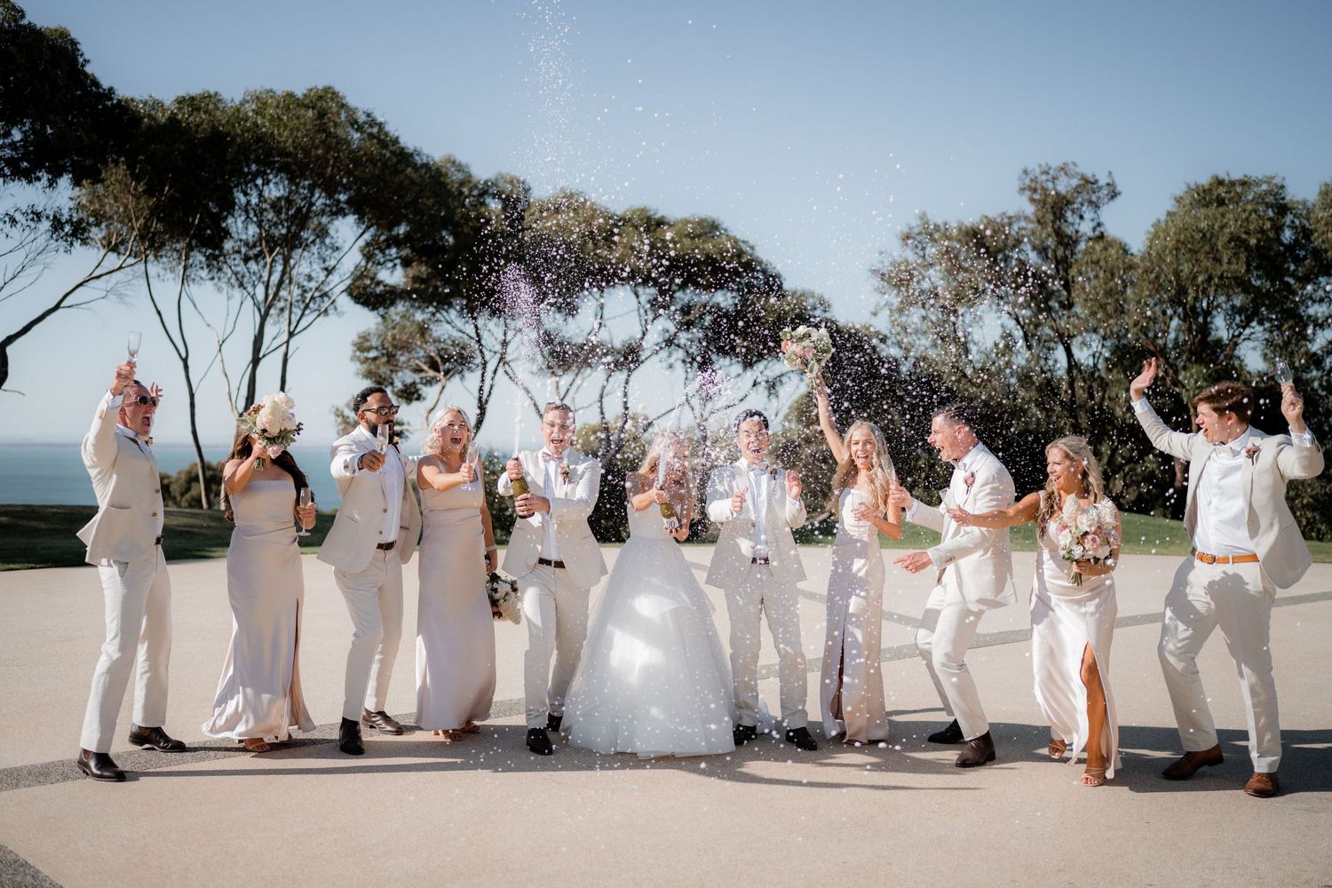 Wedding party in all white attire poses together outdoors under trees.