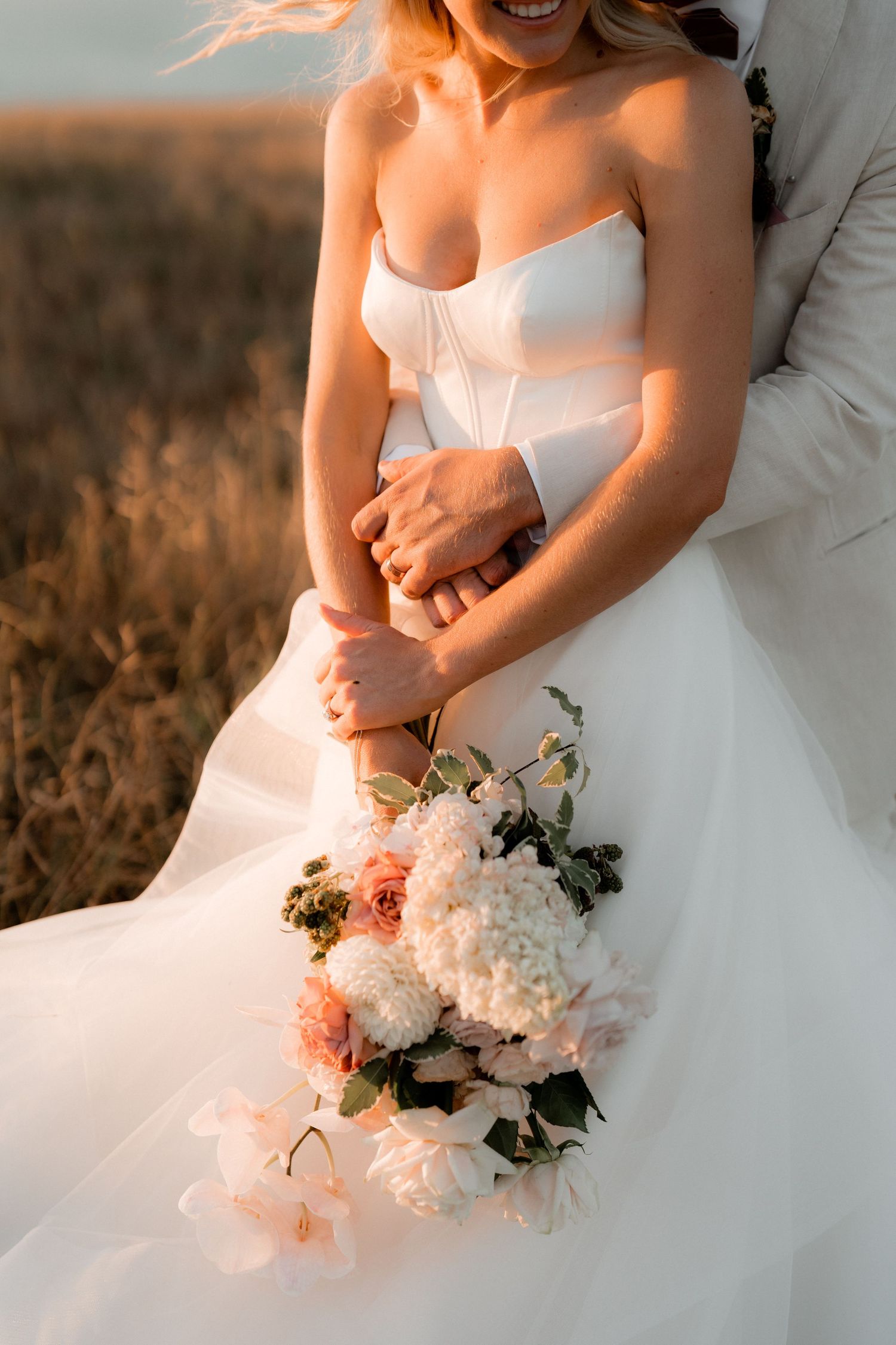 A bride in a white wedding gown holds a delicate bouquet of pink and white flowers in natural sunlight.