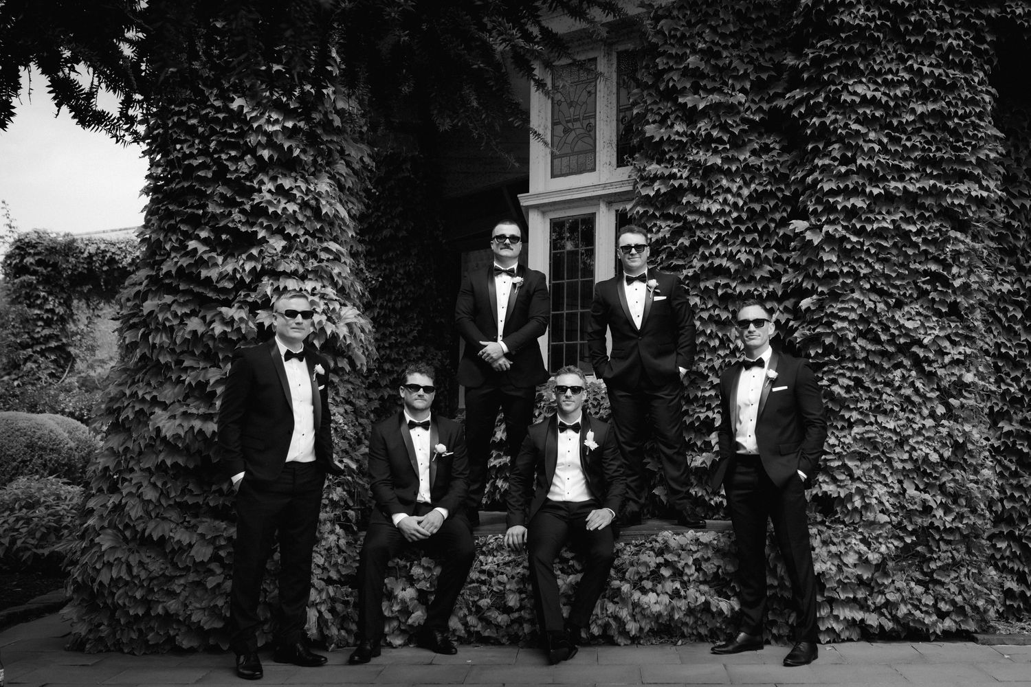 A group of formally dressed individuals in tuxedos pose in front of an ivy-covered building in a black and white photo.