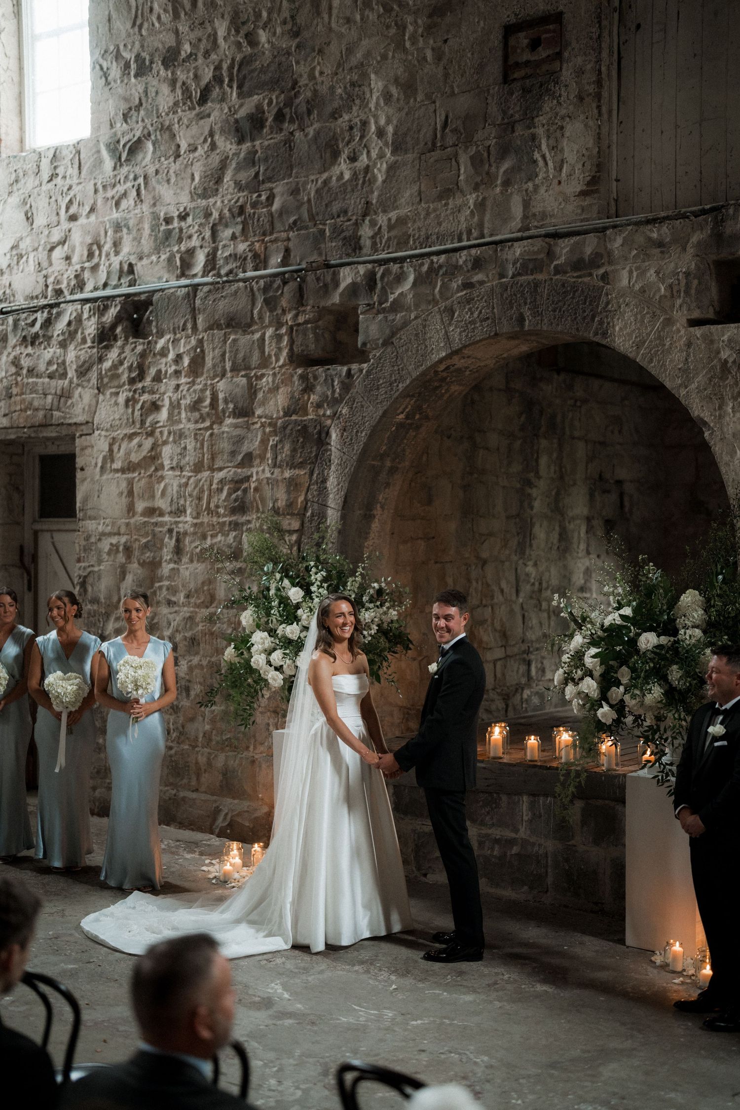 A wedding ceremony in a rustic stone cellar decorated with white flowers and candlelight.