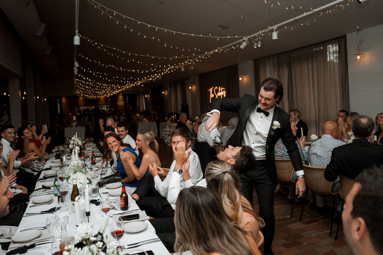 Wedding reception guests seated at long tables decorated with white linens and floral centerpieces under string lights.