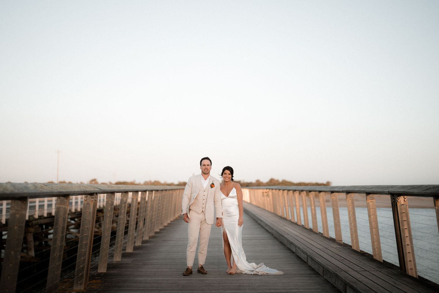 Couple walking together on a long wooden pier at sunset in light colored formal attire.