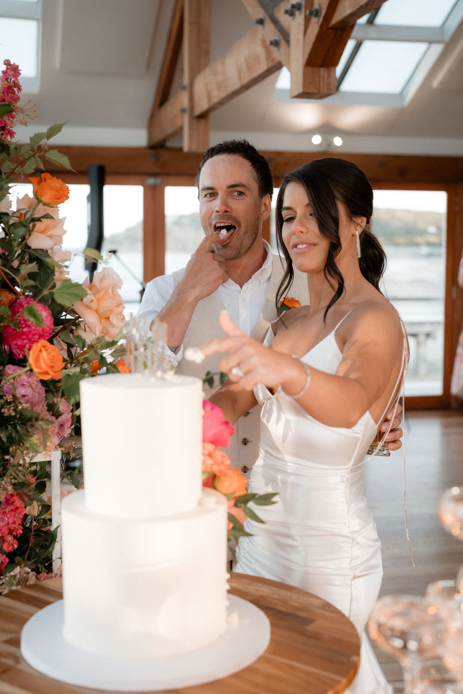 Wedding cake cutting moment in bright indoor venue with floral decorations.