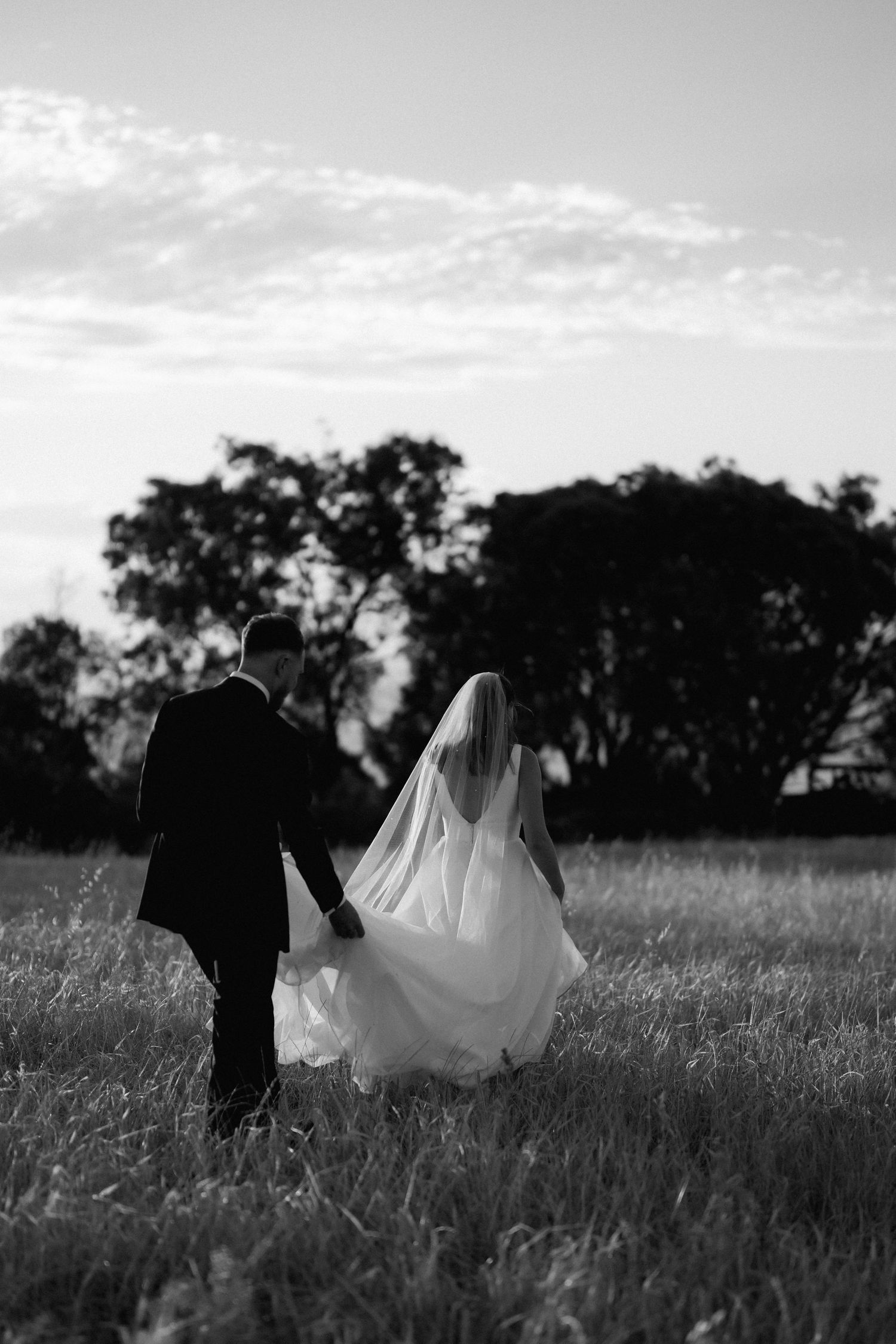 Black and white photo of newlyweds walking hand in hand through a field at dusk.