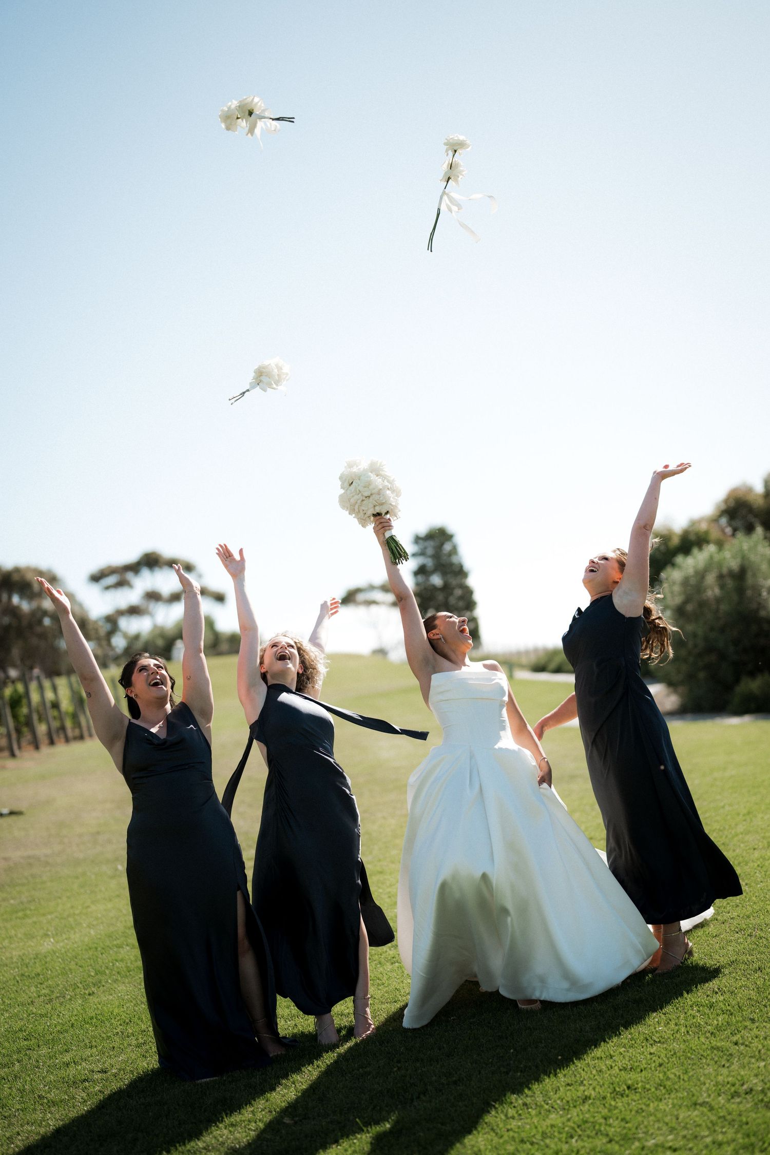 Wedding party members toss white flowers into the air while sitting on grass under blue sky.