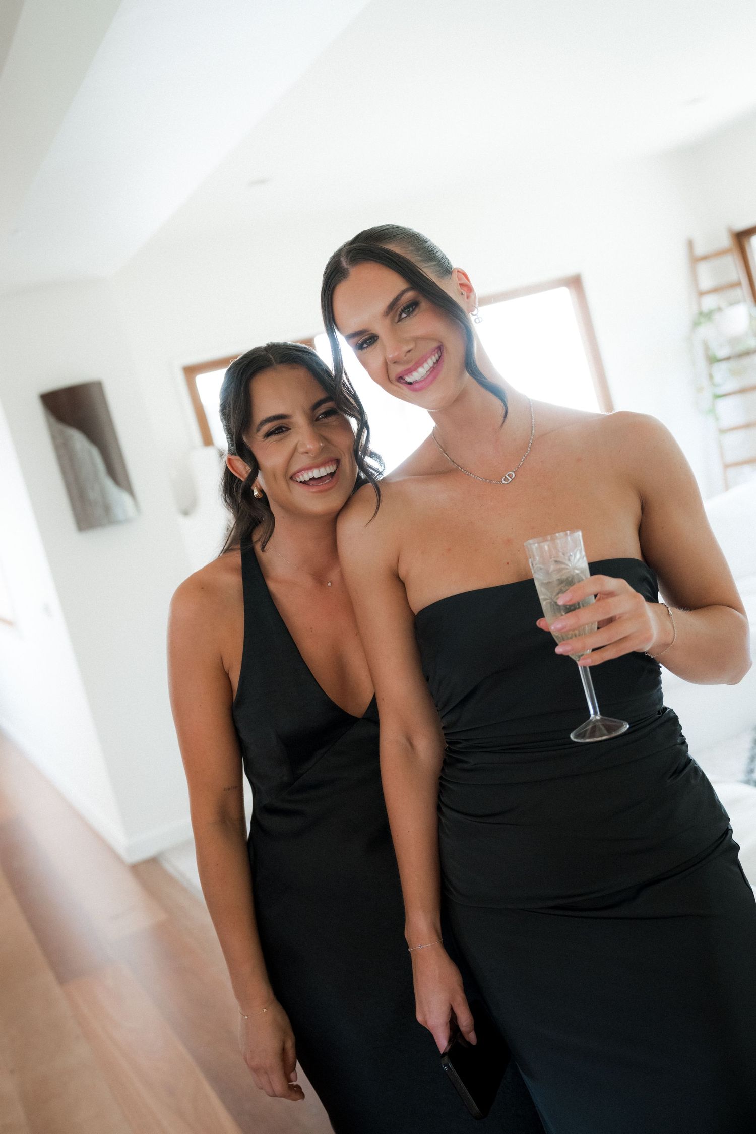 Wedding party members in black dresses sharing champagne and laughter in a bright room.