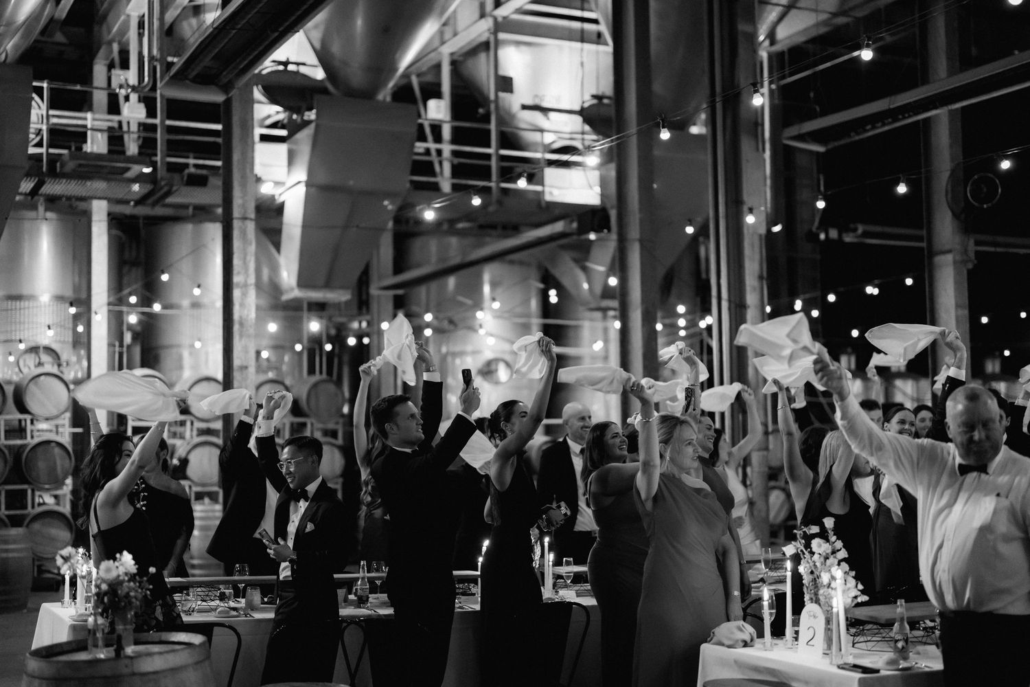 Wedding reception guests raise their hands in celebration at an industrial-style venue.