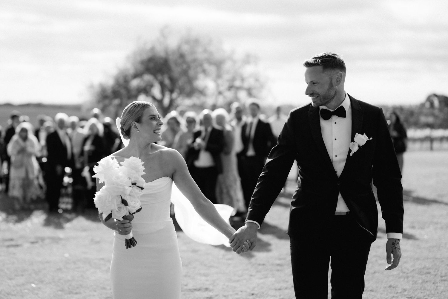 A newlywed couple walks hand in hand with guests visible in the background.