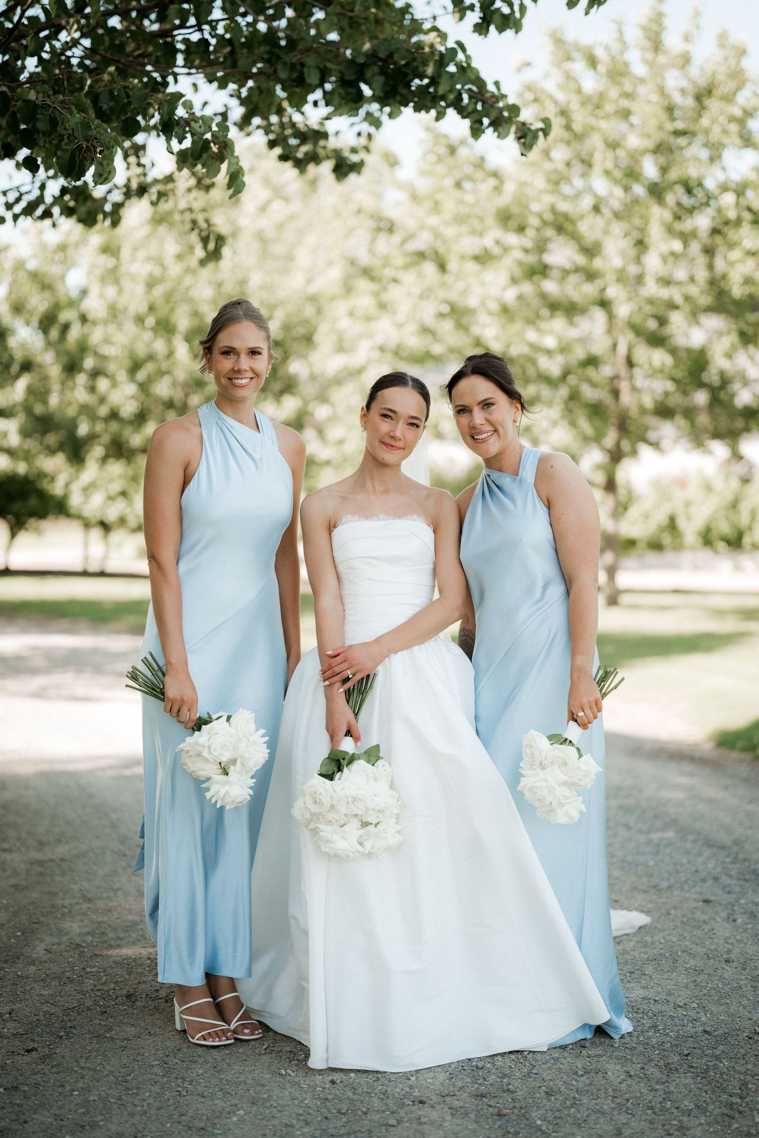 Bridesmaids in light blue dresses pose with the bride on a gravel path outdoors.