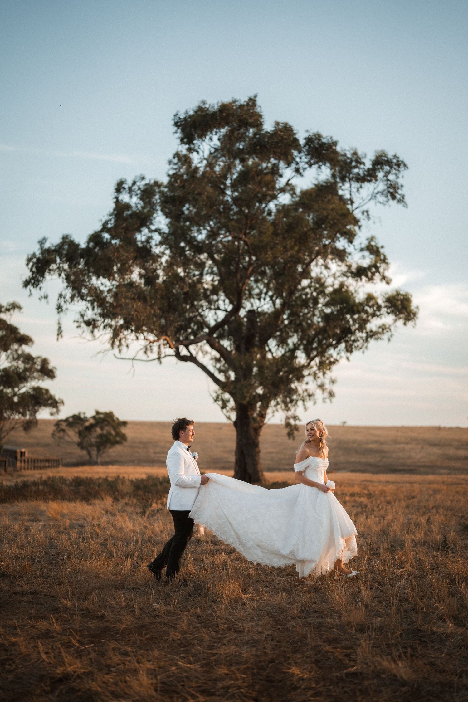 Bride and groom dancing in golden field with rustic tree backdrop at sunset.