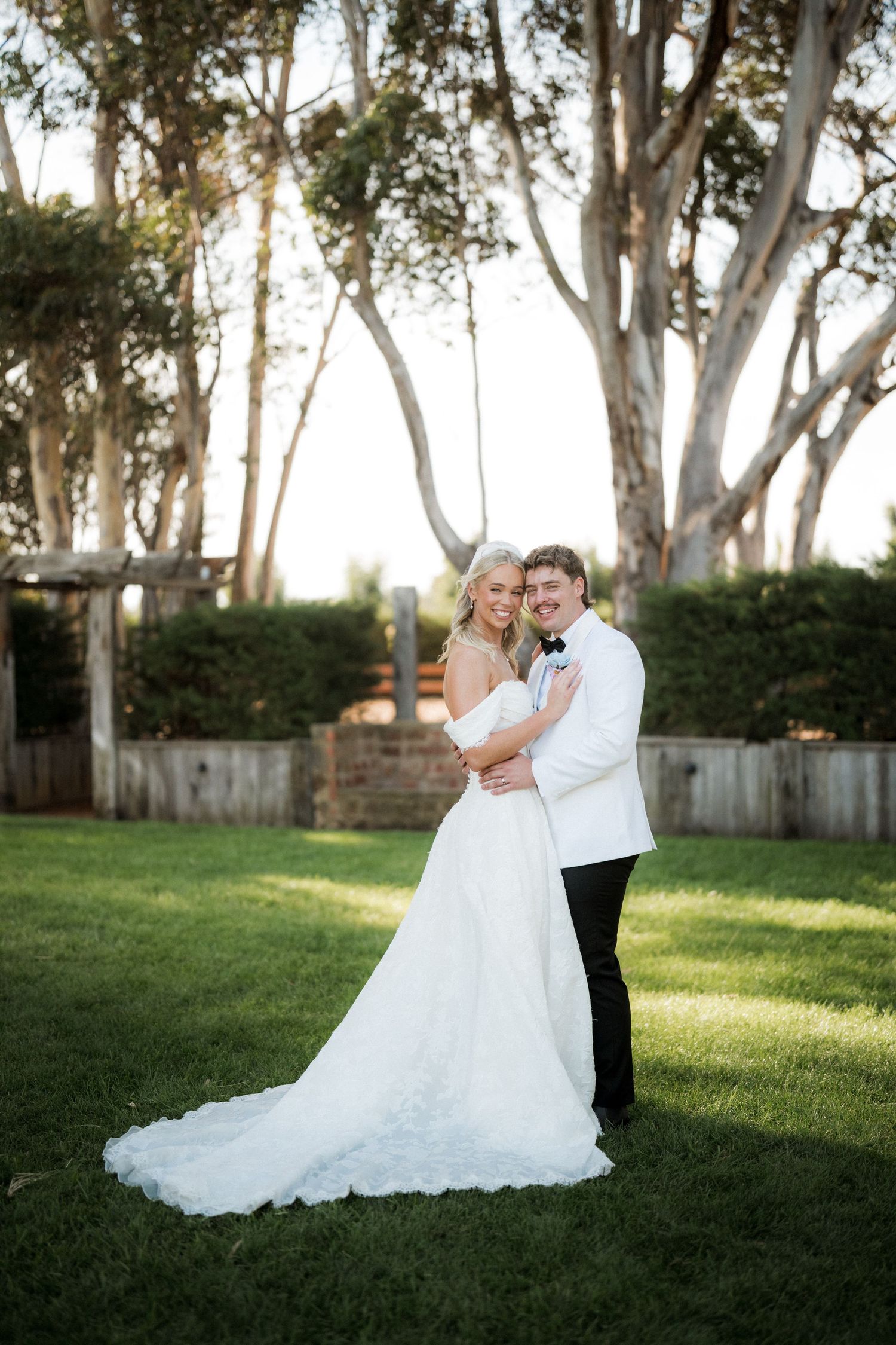 Bride and groom embracing on green lawn at golden hour with tall trees in background.