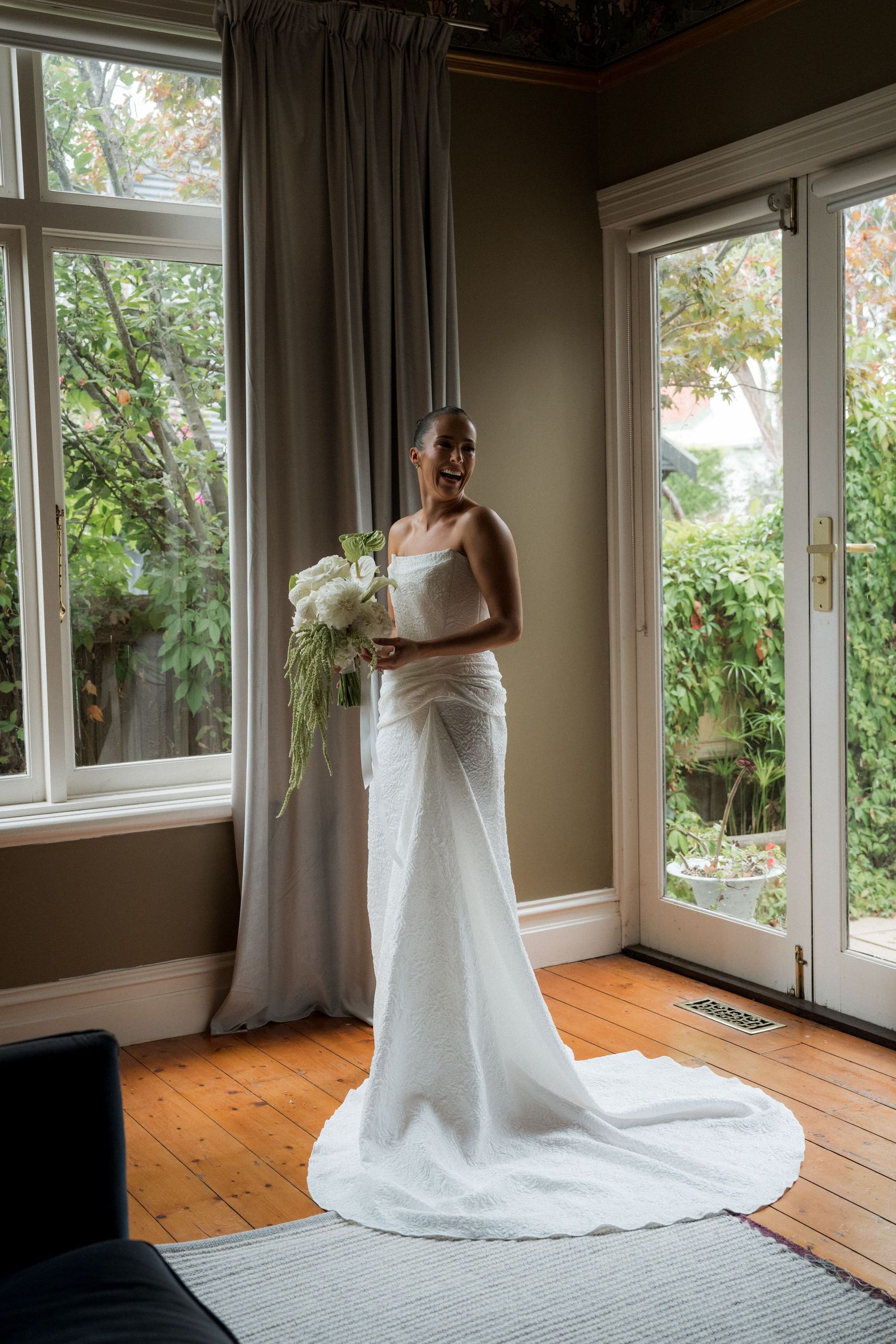 Smiling bride in a white satin gown with train holds a cascading bouquet, standing by large windows in an elegant room.