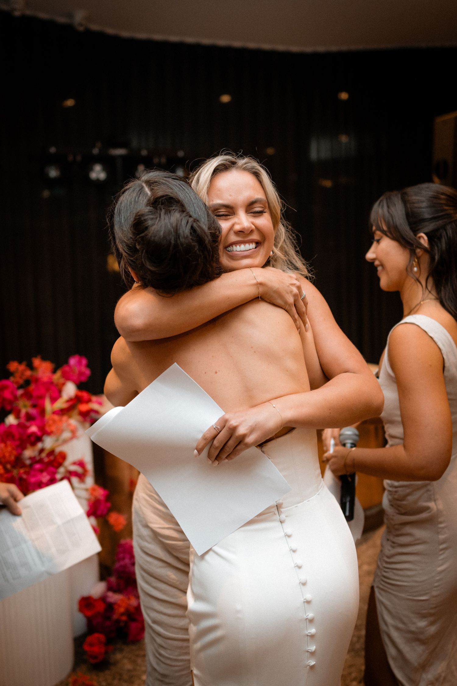 Multiple women in neutral color dresses hug and embrace during an emotional moment at a wedding reception.