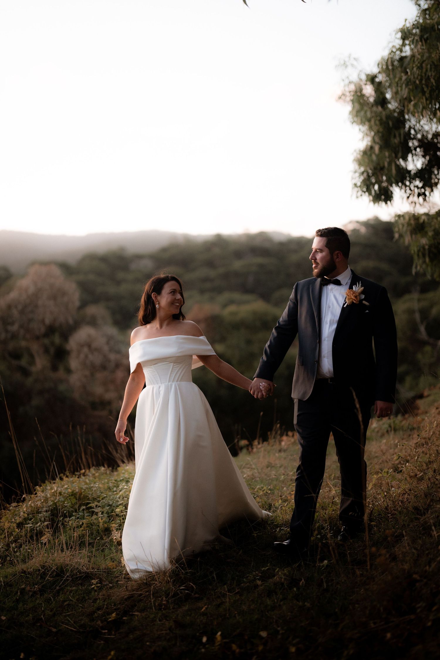 A wedding couple shares a joyful moment together on a hillside with trees and mountains in the background.