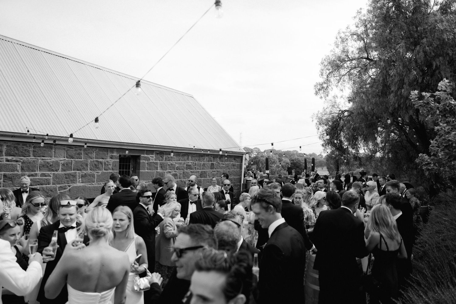 Black and white photo showing a crowd of wedding guests gathered outside a reception venue on a sunny day.