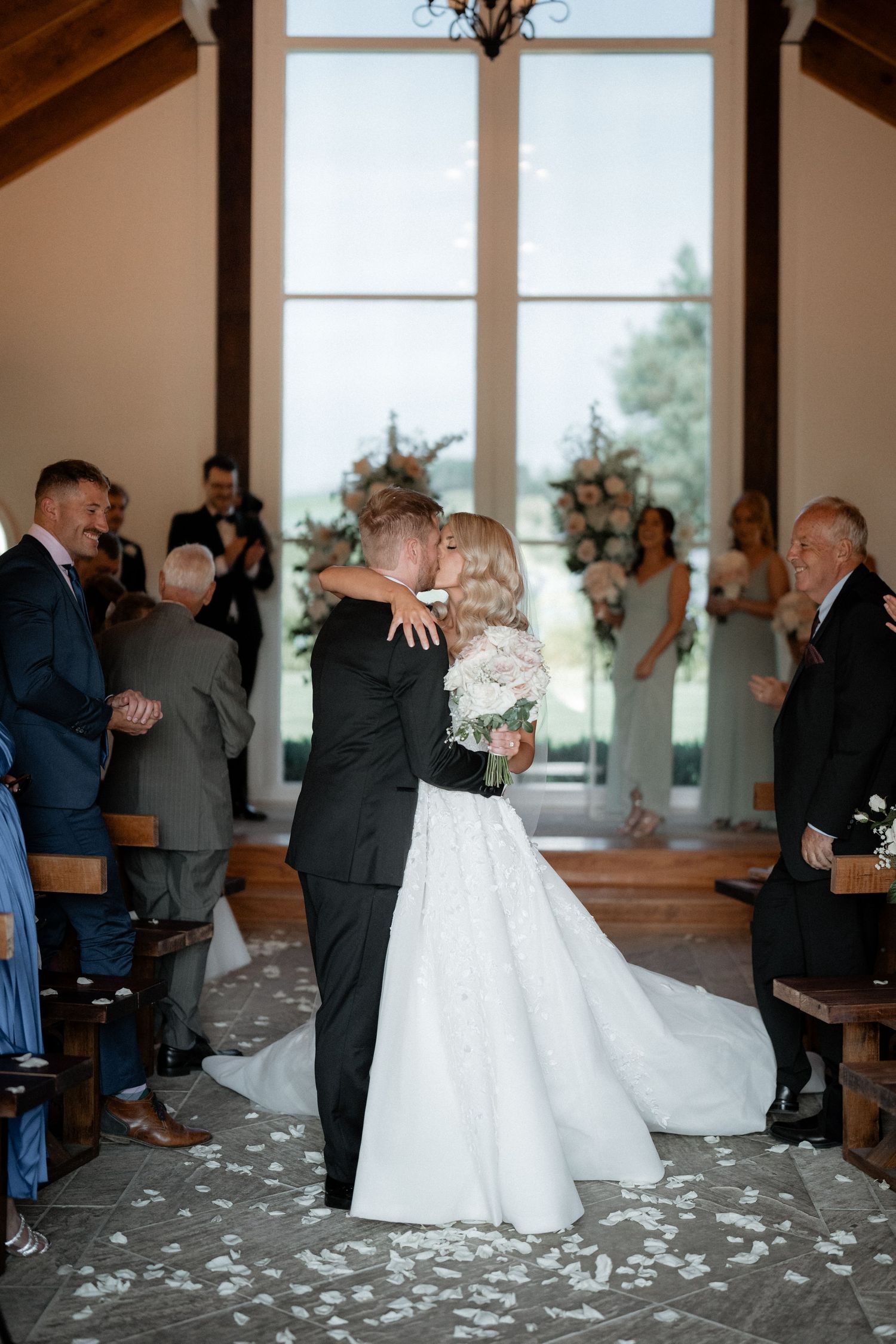 A romantic wedding ceremony kiss in a bright chapel with scattered flower petals on the floor.