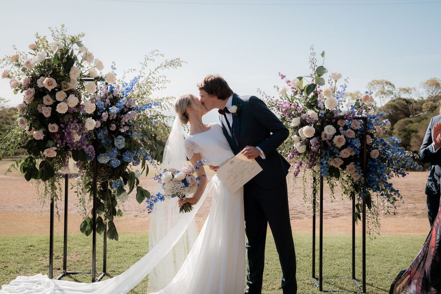 A romantic outdoor wedding ceremony with a couple sharing a kiss under floral arches with pink and blush roses.