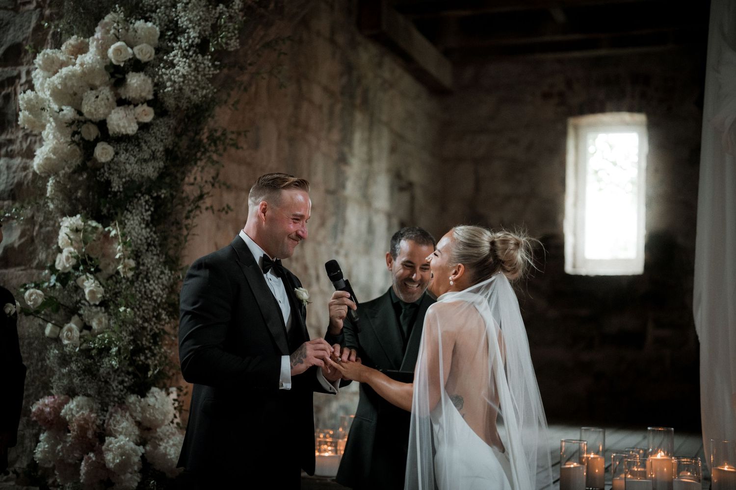 A romantic wedding ceremony takes place in a rustic stone venue decorated with white floral arrangements and candlelight.