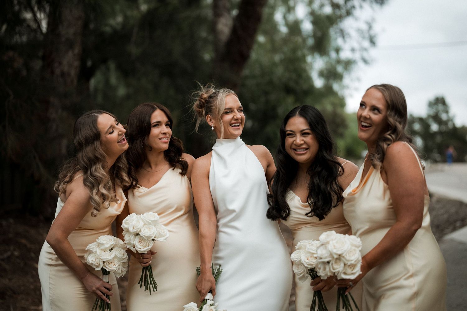 A bride and bridesmaids in neutral dresses holding white bouquets pose together outdoors for a wedding photo.