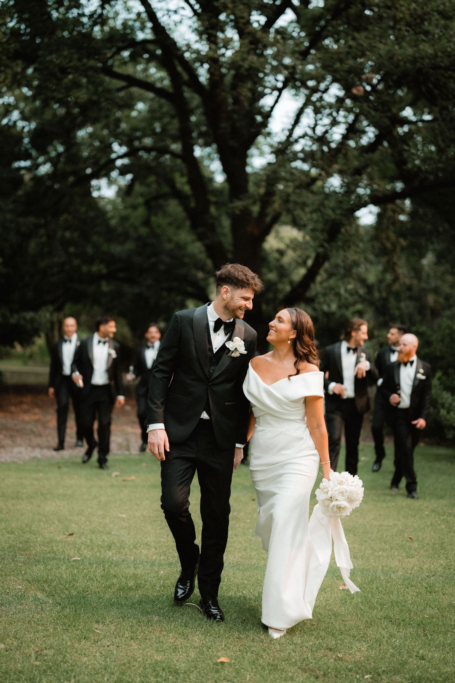 A romantic outdoor wedding scene with newlyweds walking on grass while groomsmen follow behind under tall trees.