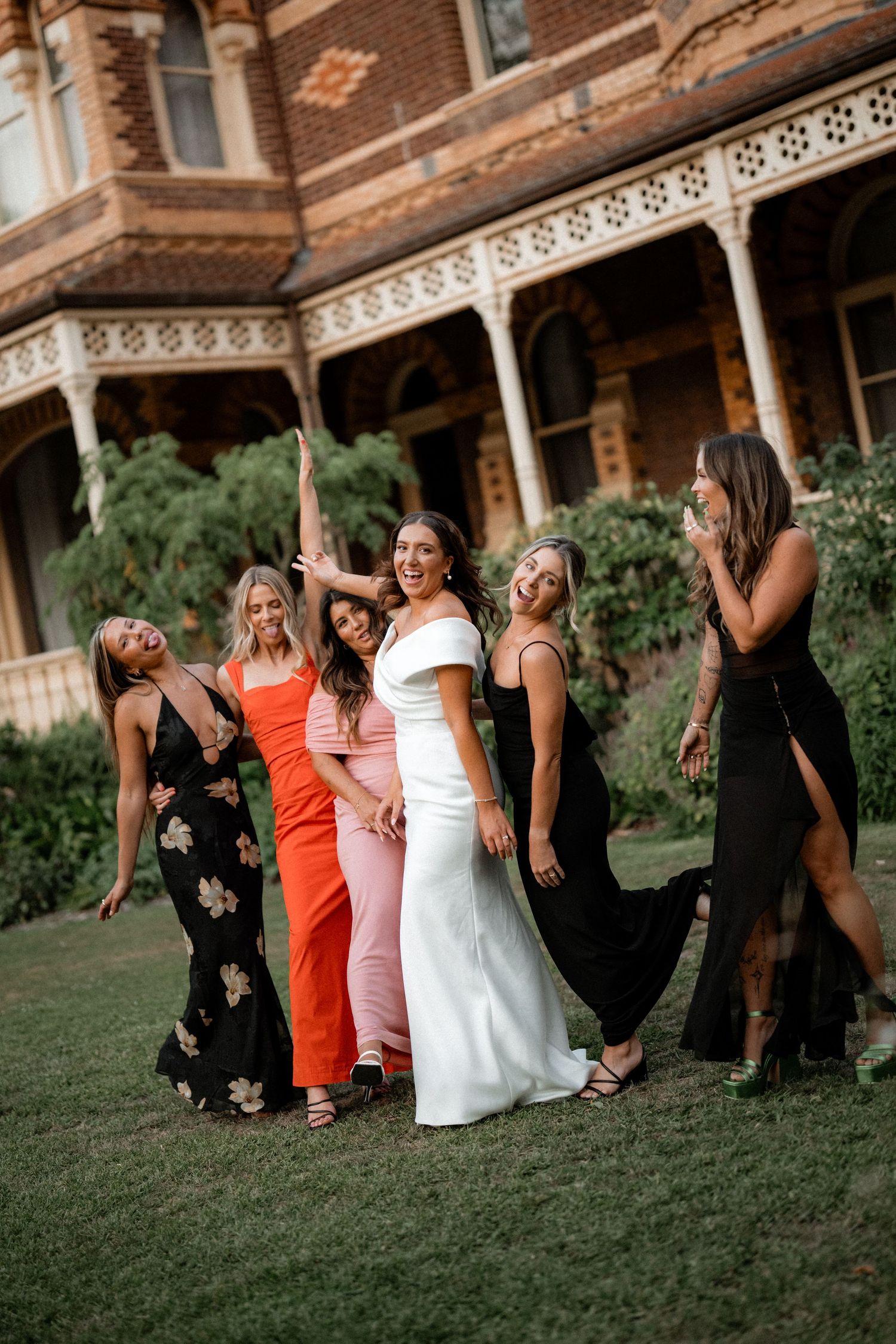 A group in formal dresses laughs and poses together on a lawn in front of a historic brick building with ornate trim.