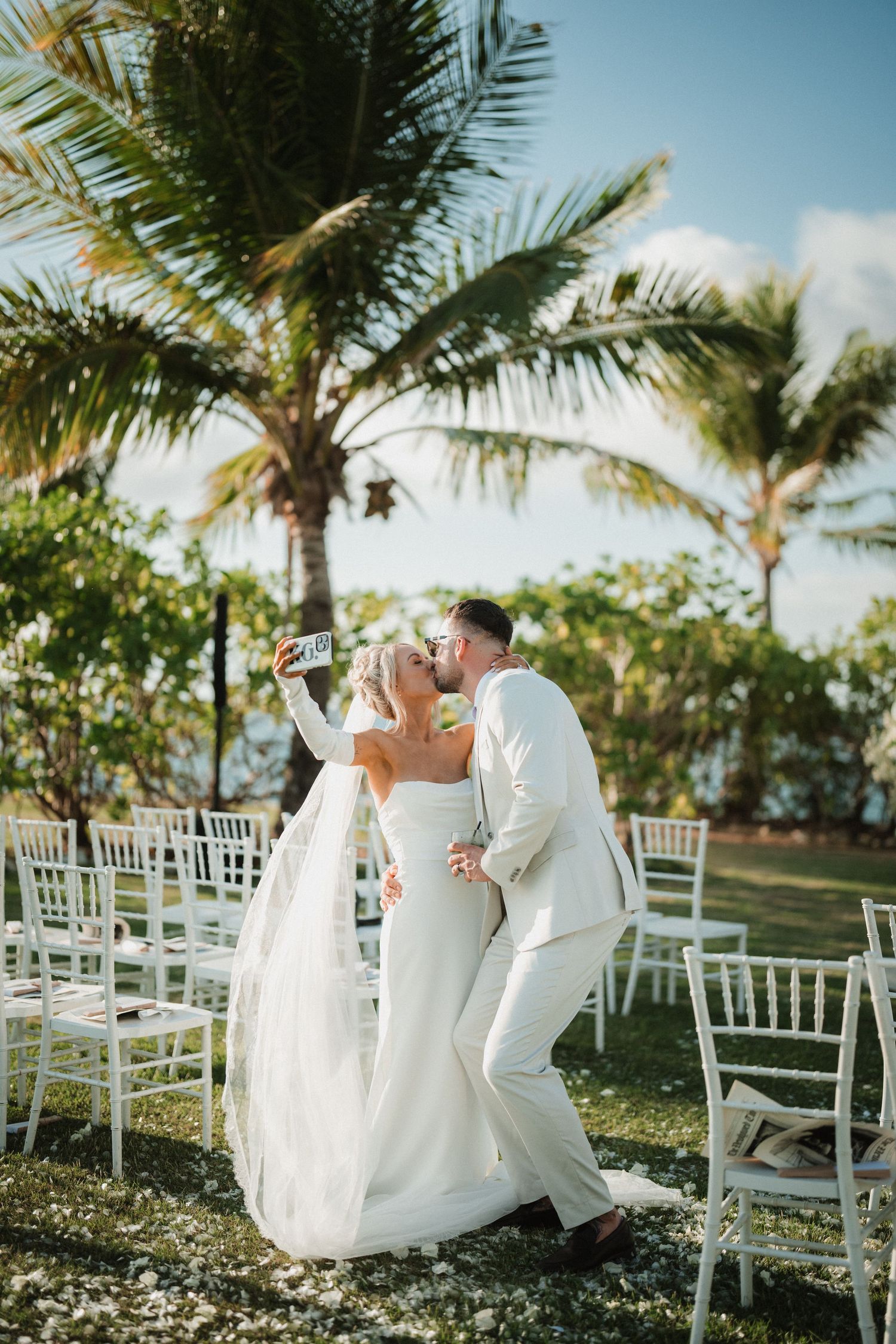 A romantic wedding ceremony setup under palm trees with white chairs on a lush tropical lawn.