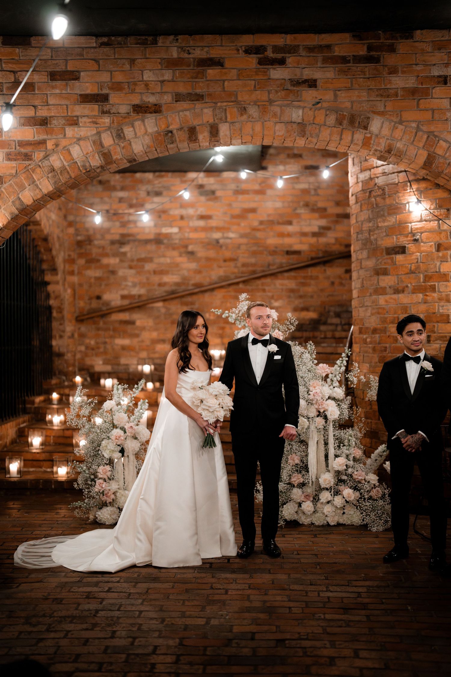 A romantic wedding ceremony under a rustic brick archway decorated with white flowers and twinkling lights.