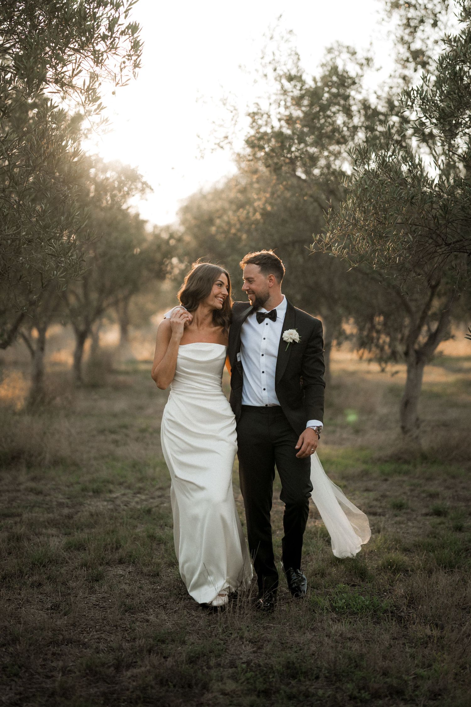 Newlyweds walk intimately through an olive grove at sunset in formal wedding attire.