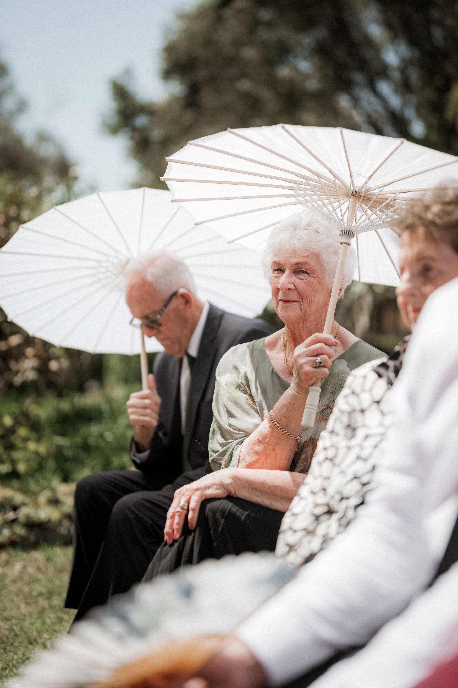 Wedding guests holding white parasols while sitting outdoors during a sunny garden ceremony.