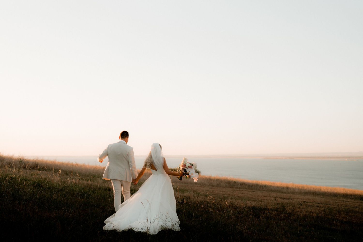 Couple in wedding attire holds hands while walking along a scenic coastal hillside at sunset.