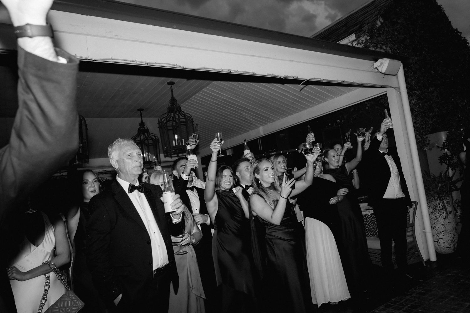 Black and white photo of formally dressed guests celebrating with raised arms under a covered outdoor space.