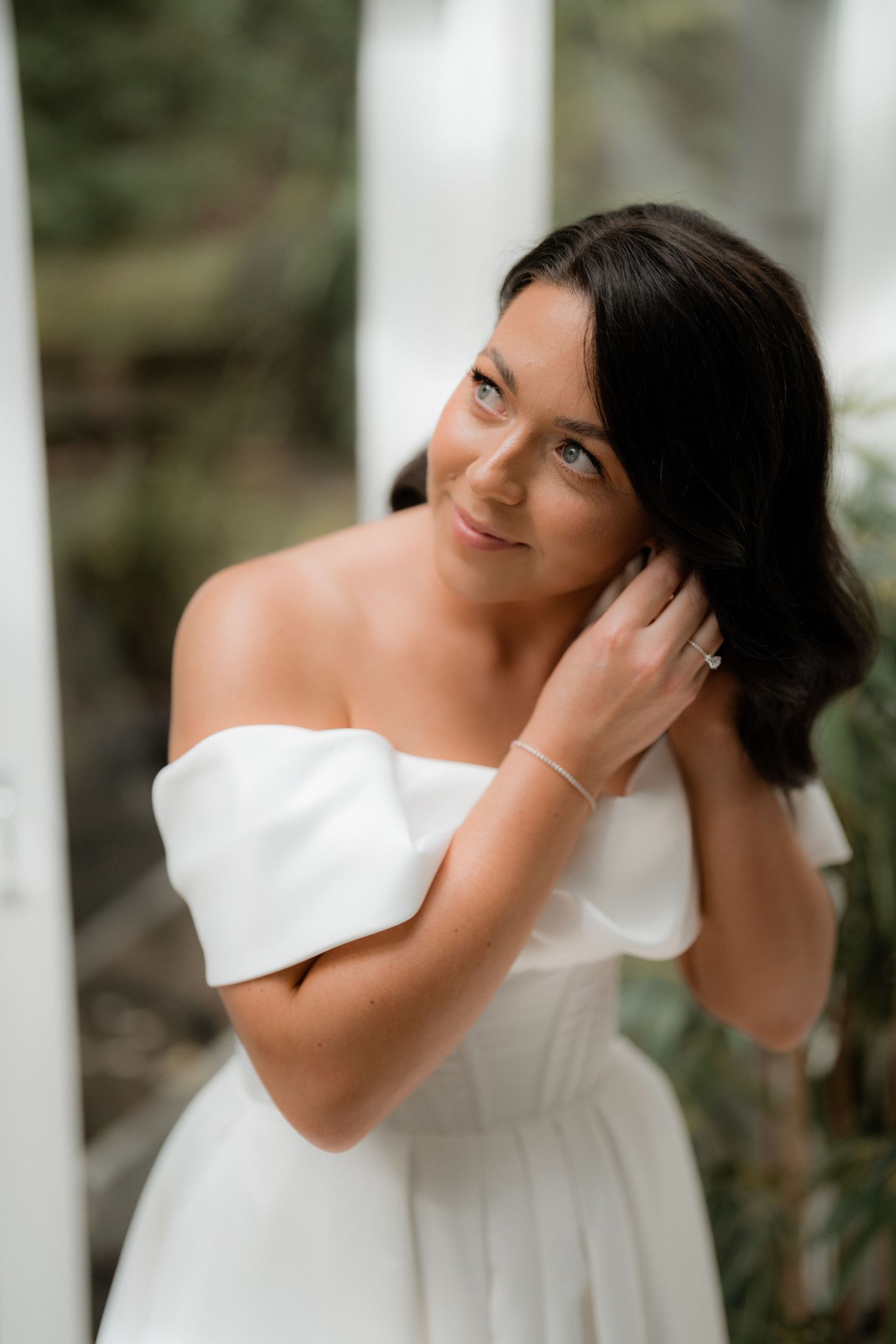 A bride in an elegant white strapless dress smiles while gently touching her dark hair in an outdoor portrait.