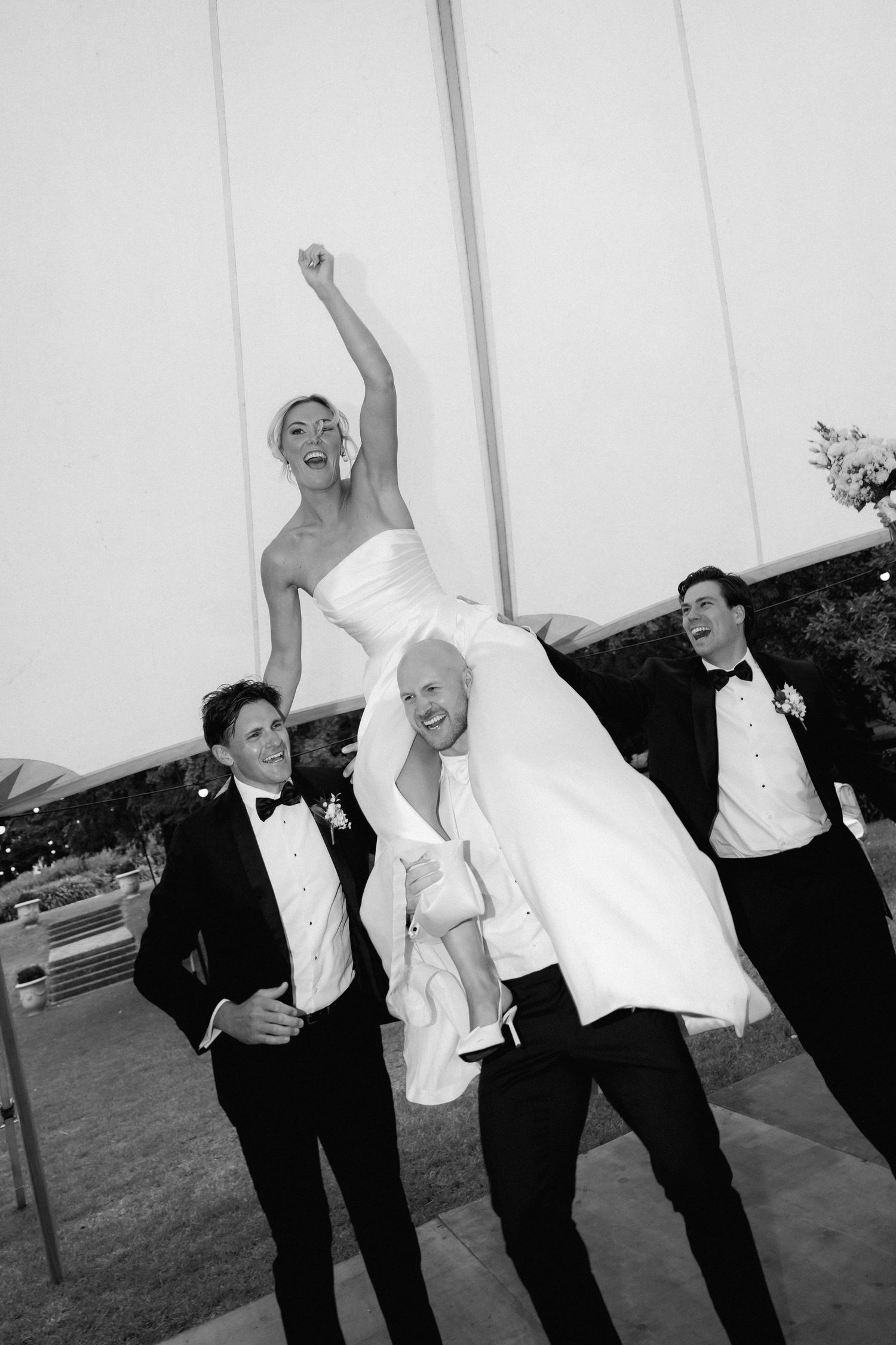 A black and white candid photo shows wedding guests lifting and carrying a bride on the dance floor.