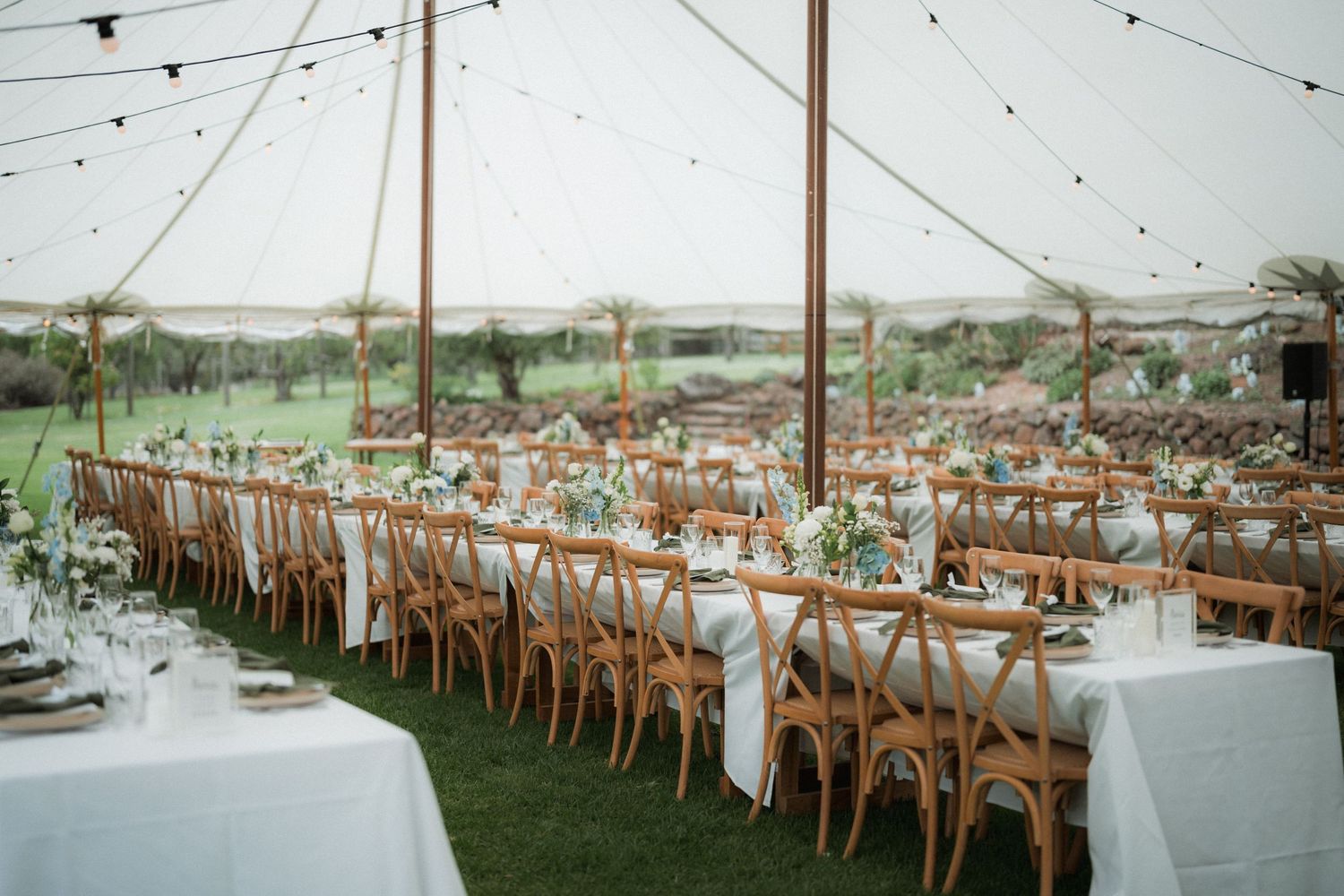 Elegant outdoor wedding reception setup with wooden cross-back chairs and white linens under a tent canopy.