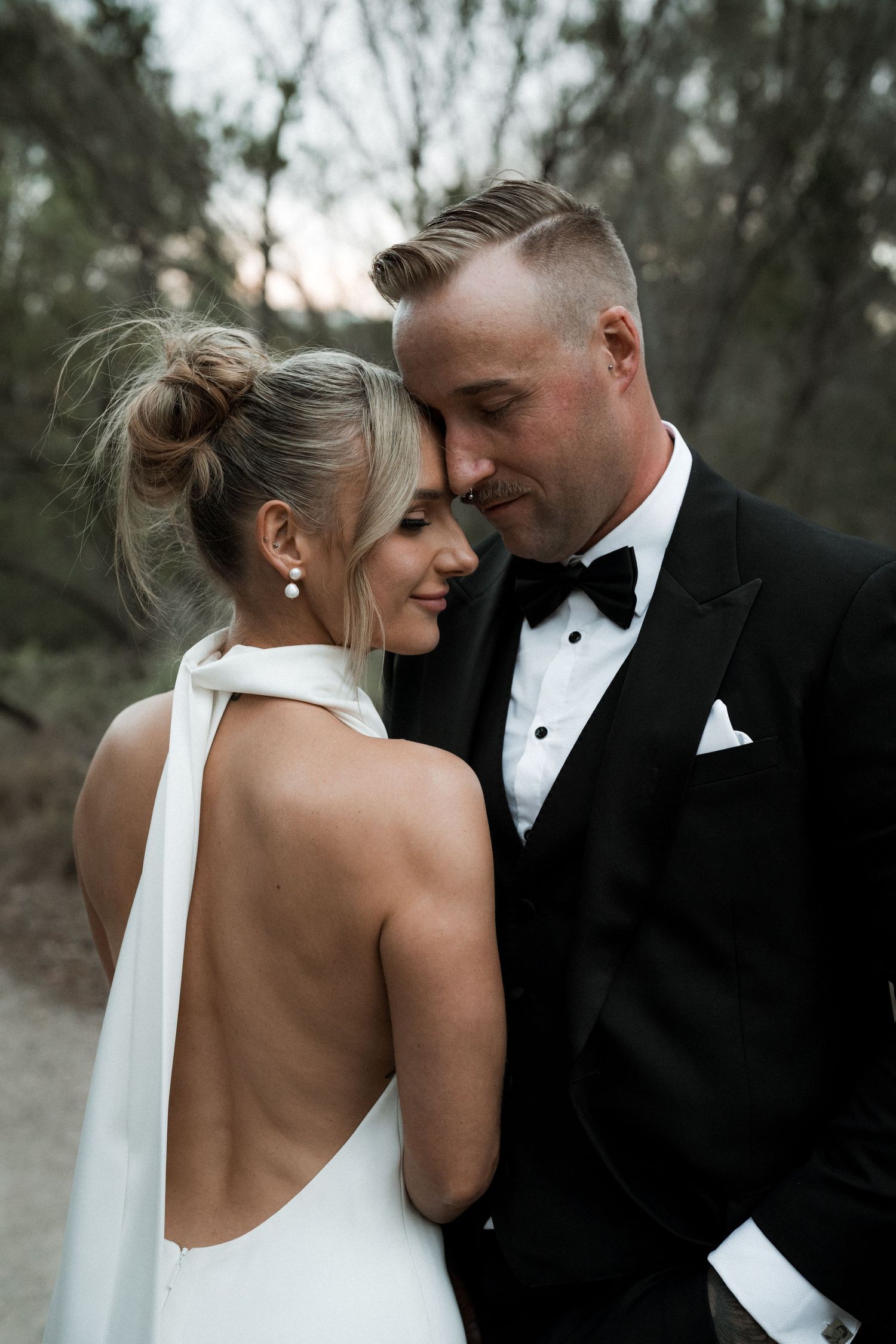 Intimate wedding portrait showing backless white gown and classic black tuxedo in moody outdoor setting at dusk.