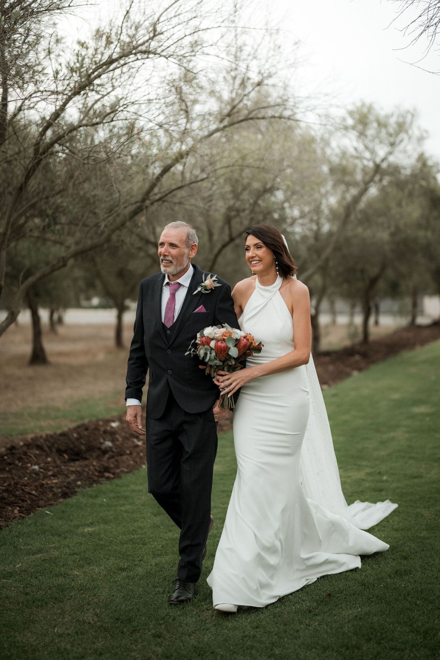 A bride in a fitted white halter dress and her escort walk together on a grassy path surrounded by bare winter trees.