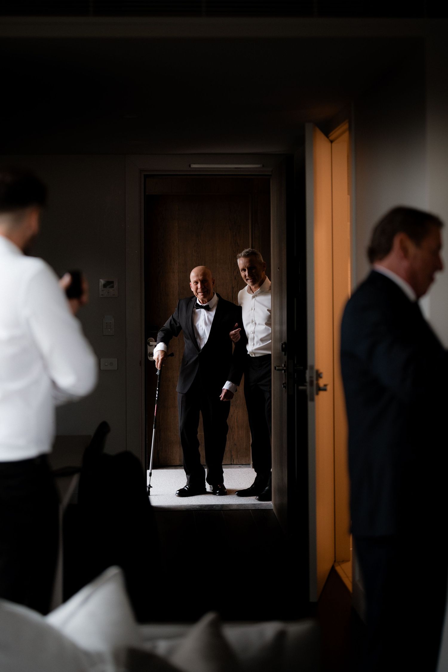 Two figures stand in a doorway while others prepare in a dimly lit hallway before an event.