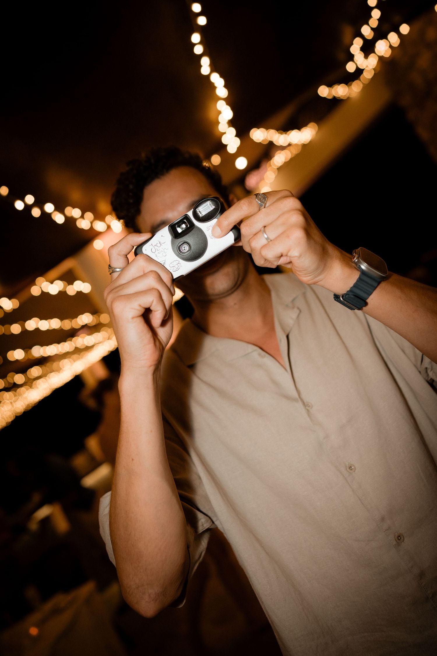 Person taking a photo with vintage camera against blurred string lights creating a bokeh effect at night.