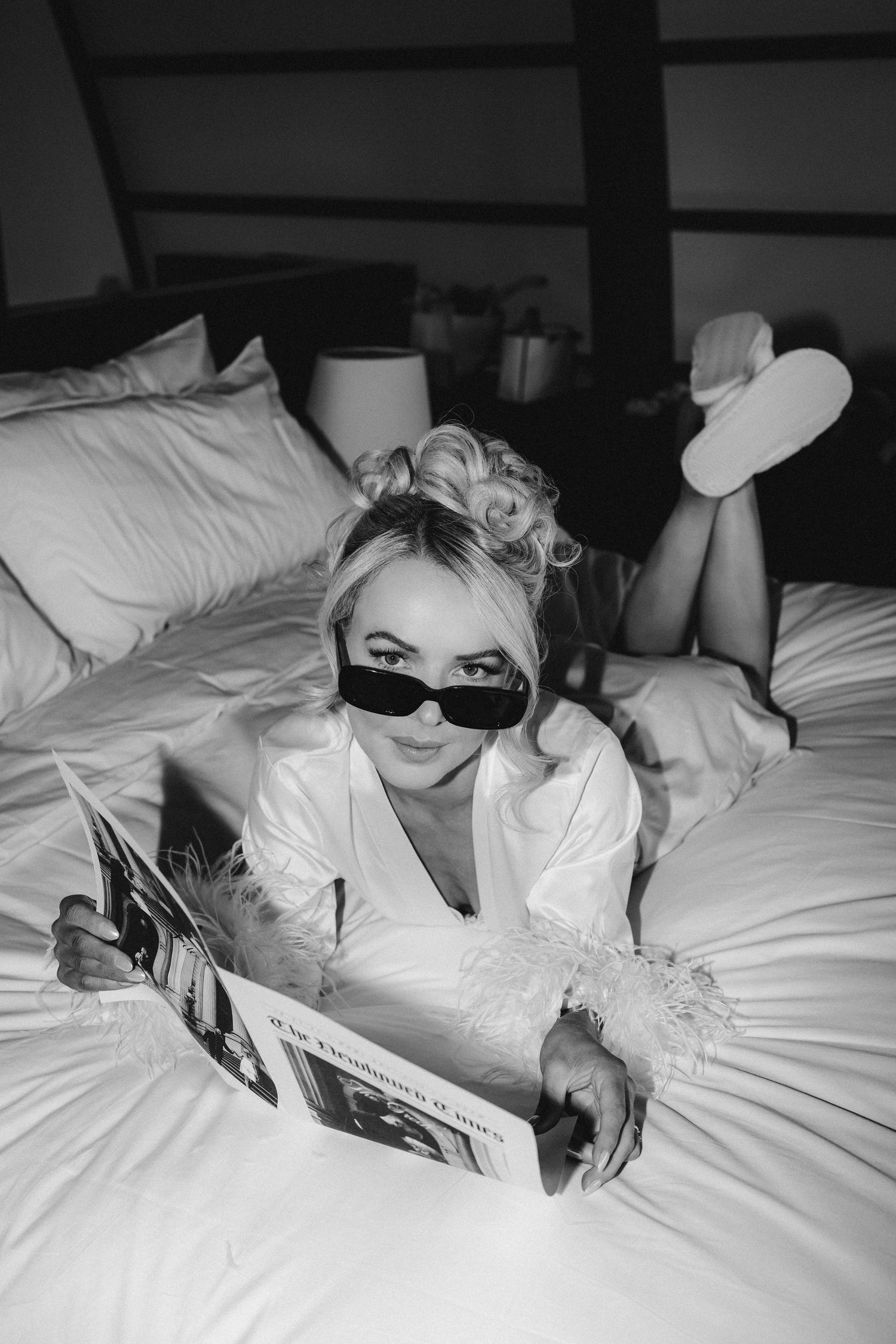 A stylish black and white boudoir photo of a woman in sunglasses and white robe lounging on bed with a magazine.
