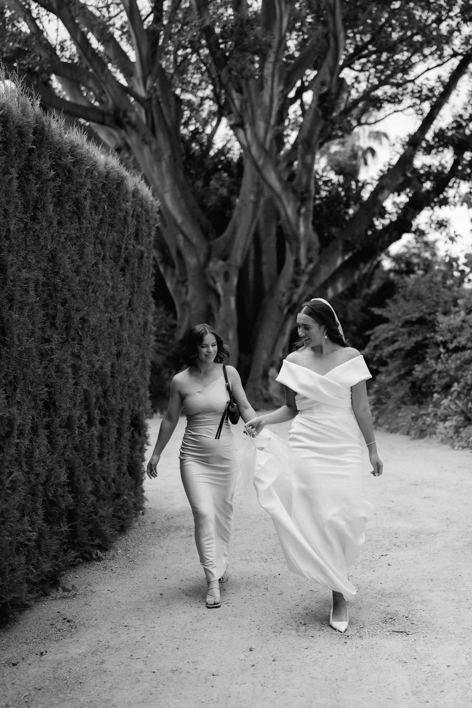 Two women walking together on a garden path, one wearing a fitted gown and one in an elegant white dress.
