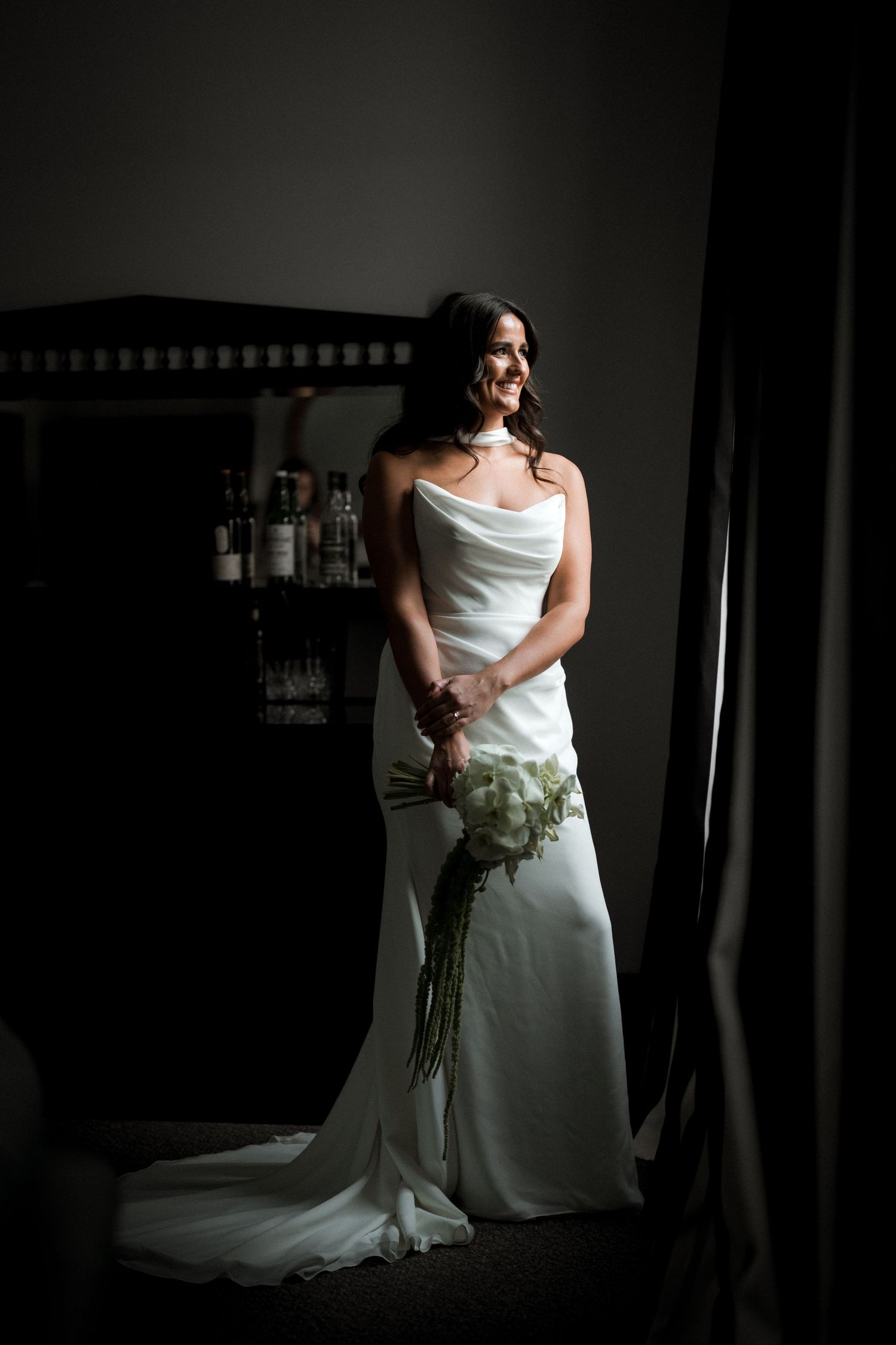 A figure in an elegant white wedding gown stands by a window in dramatic lighting holding a white floral bouquet.